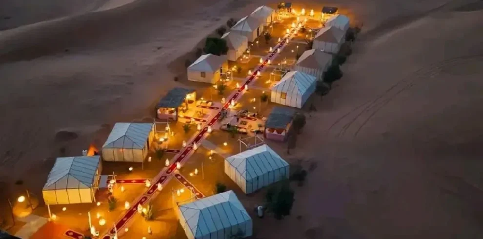 Prompt: Luxury desert bivouac in Merzouga Sahara, Morocco — traditional Berber tents, golden sand dunes, warm sunset light, campfire with carpets and lanterns, cozy desert atmosphere.