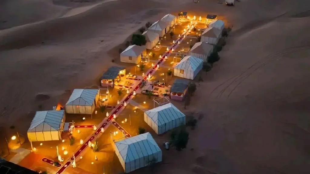 Prompt: Luxury desert bivouac in Merzouga Sahara, Morocco — traditional Berber tents, golden sand dunes, warm sunset light, campfire with carpets and lanterns, cozy desert atmosphere.